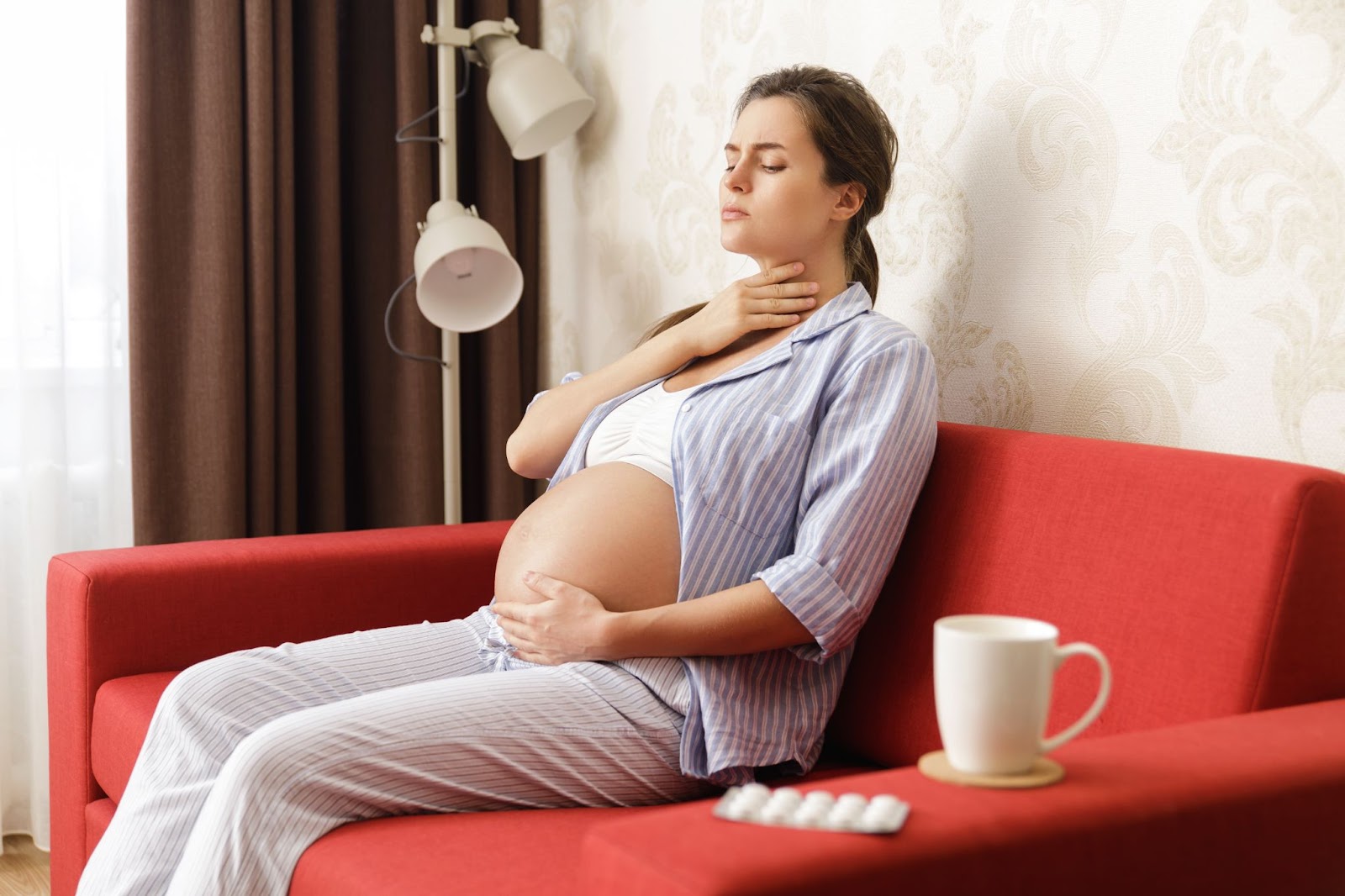 Pregnant woman with sore throat sits on her couch with a cup of hot tea and lozenges.